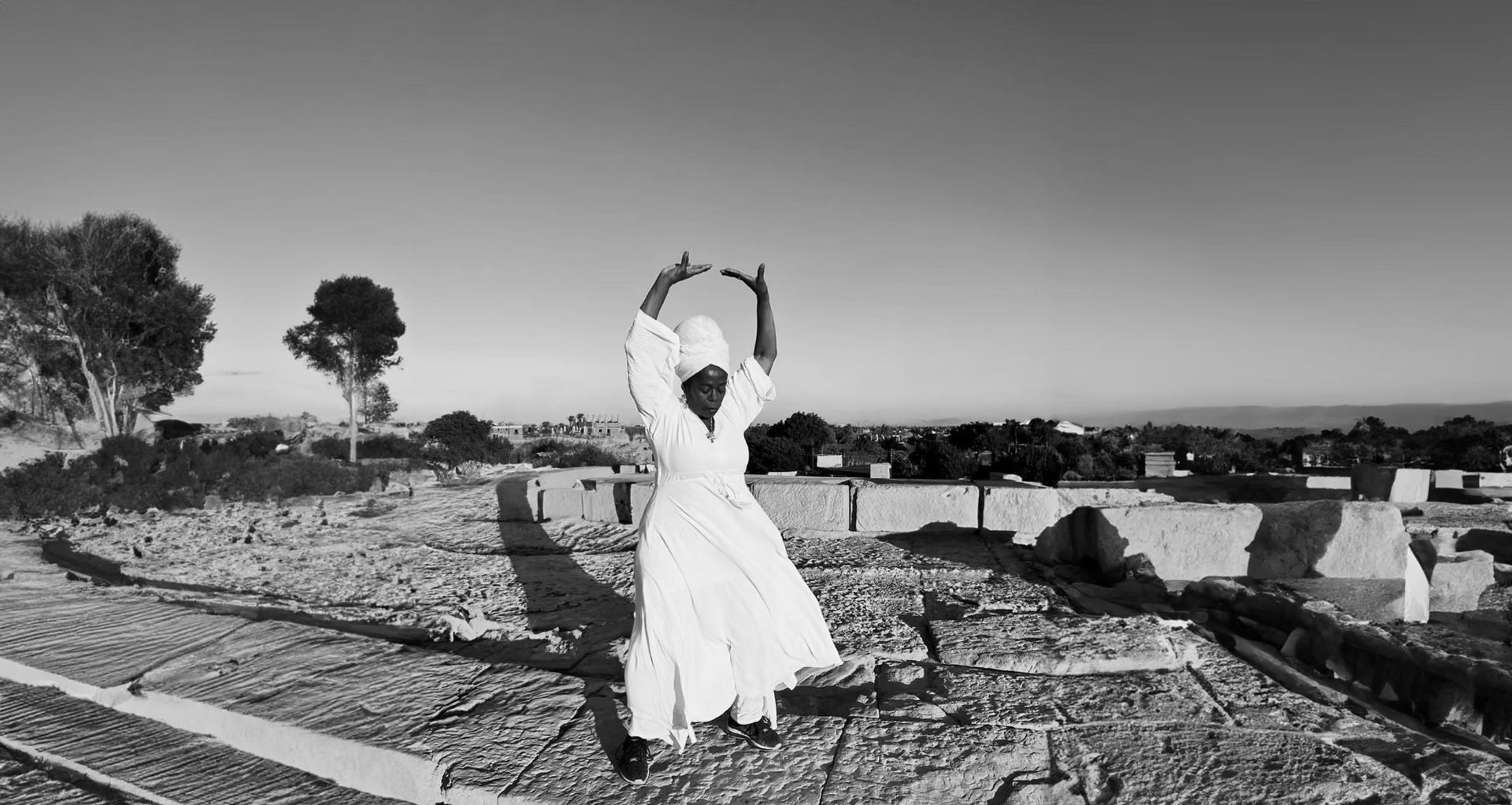 Sandra Golding dancing in a flowing white dress and headwrap on a sunlit stone landscape